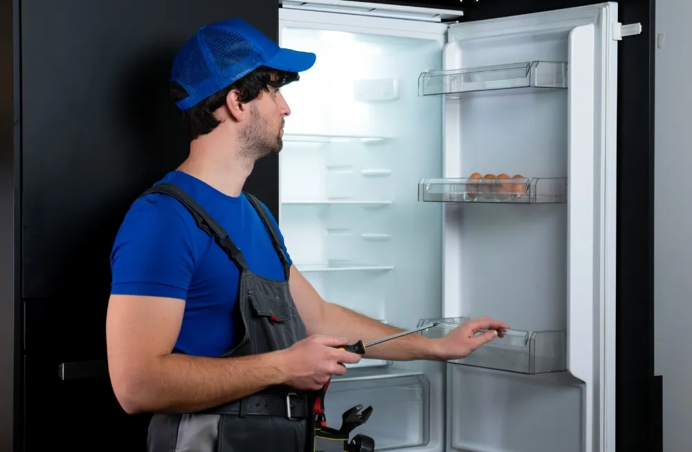 A technician wearing a blue shirt and cap is inspecting an empty refrigerator, using a screwdriver to check the appliance.