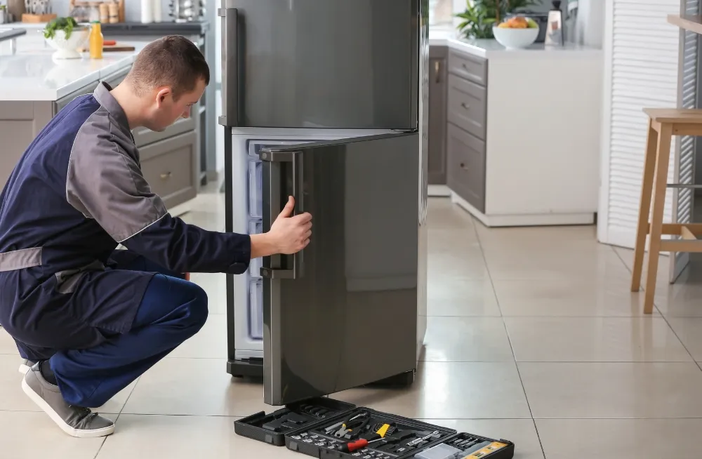 Technician in a blue uniform repairing a modern double-door refrigerator in a kitchen, with a toolbox and tools nearby.