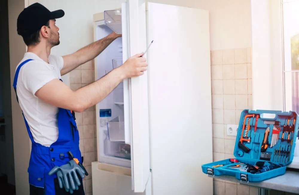 Technician in Dubai repairing a white refrigerator with tools in a kitchen.