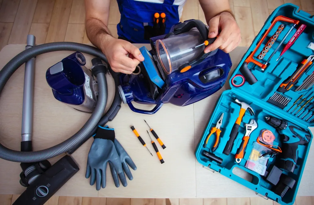 Person repairing a blue vacuum cleaner with tools on a wooden table, showcasing maintenance and repair work.