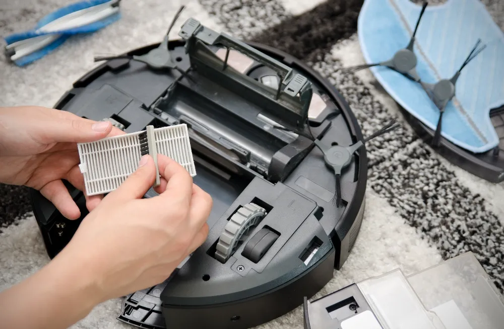 Hands replacing a filter on an upside-down robotic vacuum cleaner, surrounded by spare parts on a patterned carpet. Vacuum Cleaner Repair Dubai