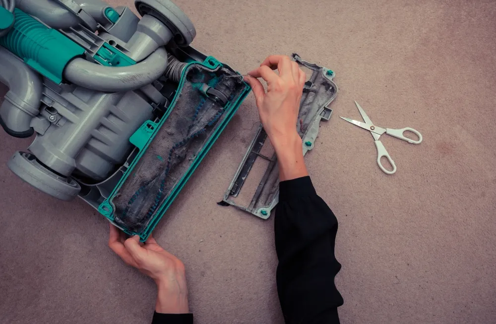 Person cleaning a vacuum cleaner's tangled brush roll with scissors on a carpeted surface. Vacuum Cleaner Repair Dubai - PrimeProTechnical