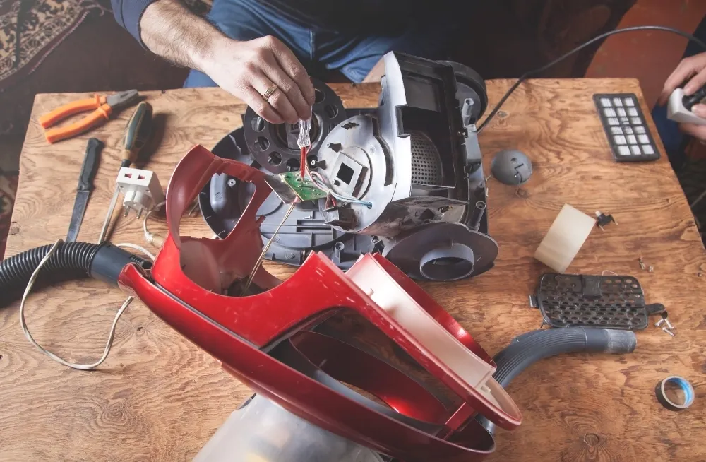 Hands repairing a disassembled vacuum cleaner on a wooden table, focusing on internal components and circuit board.